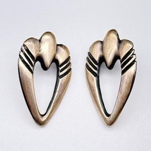 Puede incluir: Un par de pendientes plateados vintage. Cada pendiente tiene un diseño en forma de corazón con una parte superior redondeada y una base puntiaguda. El diseño incluye tres líneas horizontales a cada lado del corazón.