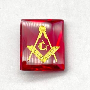 May include: A square, ruby-red Masonic emblem with a gold-colored square and compass symbol. The symbol is centered on the square, which has a slightly translucent appearance. The background is a light, neutral color.