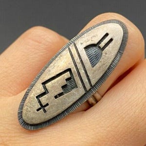May include: A silver-toned ring with an elongated oval face. The face features etched geometric designs and a textured border. The ring is worn on a finger, showcasing the intricate details and craftsmanship of the jewelry.