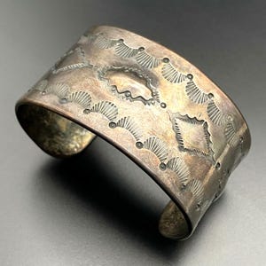 May include: A silver-toned cuff bracelet with an engraved geometric pattern. The design includes fan-shaped and diamond-shaped elements, along with small circular details. The bracelet has an aged appearance, suggesting it is vintage or antique.