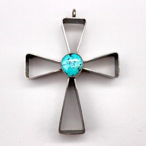 May include: A silver-toned metal cross pendant featuring an open, geometric frame design. At the center of the cross is a single, round, bright turquoise stone with dark matrix inclusions. A small metal loop is attached to the top for hanging.
