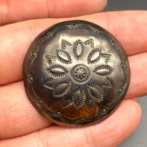 May include: A round, silver-toned metal button with an embossed floral design. The button features a central starburst surrounded by oval shapes and detailed edges. The button is held in a hand.
