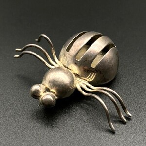 Könnte beinhalten: Silberfarbene Spinnenbrosche mit rundem Körper und segmentierten Beinen. Der Körper der Spinne hat ein gestreiftes Design, und die Augen sind kleine Kugeln. Die Brosche ist ein einzigartiges Schmuckstück.