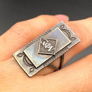 Op de afbeelding: Zilverkleurige ring met een rechthoekige voorkant met een kleinere vierkante en ruitvormige accenten. De ring heeft een uniek ontwerp met een getextureerd oppervlak en een decoratief embleem in het midden. De ring wordt aan een vinger gedragen.