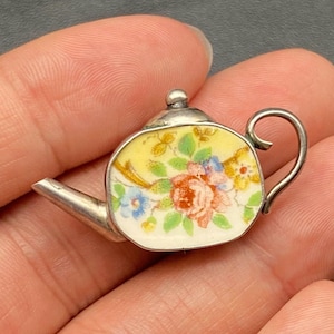 Könnte beinhalten: Ein Miniatur-Teekannen-Anhänger mit einem silberfarbenen Metallrahmen. Die Teekanne hat einen gelben Hintergrund mit einem Blumendesign in Rot-, Blau- und Grüntönen. Der Griff und der Ausguss sind ebenfalls silberfarben.
