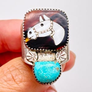 May include: A silver ring featuring a horse head design. The ring has a rectangular face with a brown and black background and a white horse head. A turquoise stone is set at the base of the ring.