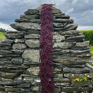 May include: A long, vertical macrame wall hanging in a deep burgundy color. The macrame design features a repeating pattern of interconnected leaf-like shapes. The piece is suspended from a natural wooden dowel, creating a rustic, bohemian aesthetic.