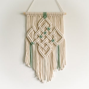 May include: A white macrame wall hanging with a geometric design featuring green accents. The hanging is made with cotton cord and has a wooden dowel at the top.