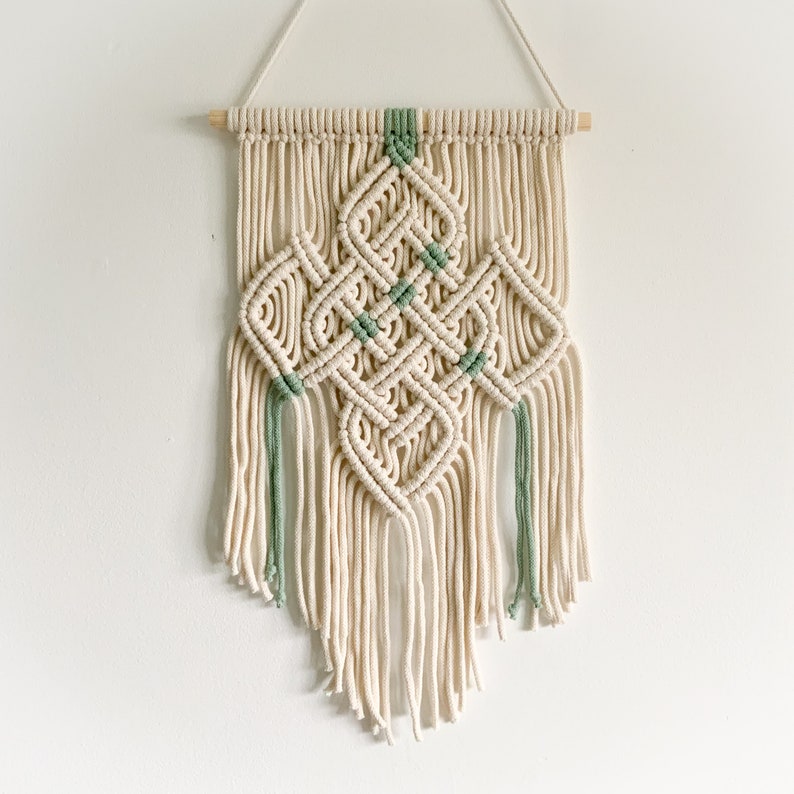 May include: A white macrame wall hanging with a geometric design featuring green accents. The hanging is made with cotton cord and has a wooden dowel at the top.