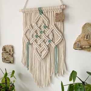 May include: A white and green macrame wall hanging with a diamond pattern and fringe. The hanging has a wooden dowel at the top and a small tag that reads "Cotton and Macrame Handmade in Poland".