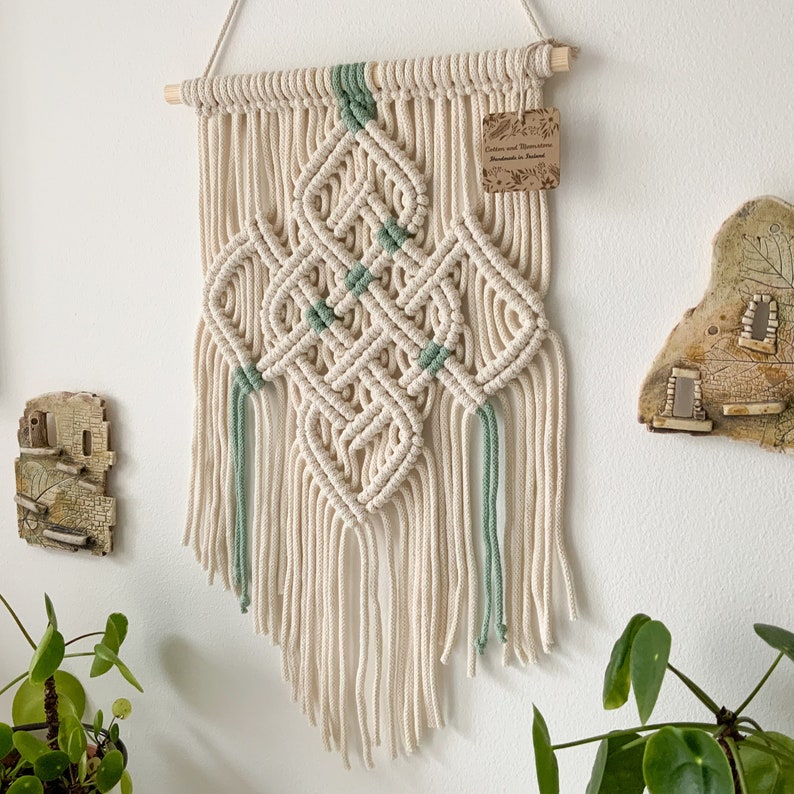 May include: A white and green macrame wall hanging with a diamond pattern and fringe. The hanging has a wooden dowel at the top and a small tag that reads "Cotton and Macrame Handmade in Poland".