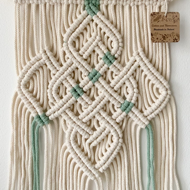 May include: A macrame wall hanging with a geometric design in cream and sage green. The hanging has a tag that reads "Cotton and Macrame Handmade in Ireland".