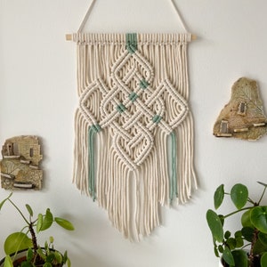 May include: A macrame wall hanging with a diamond pattern in beige and green. The hanging has a wooden dowel at the top and long fringe at the bottom.