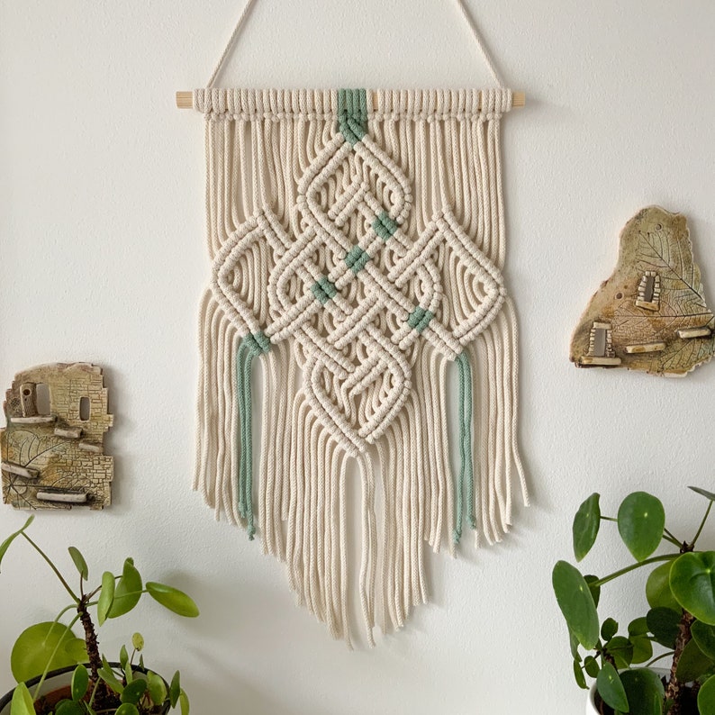 May include: A macrame wall hanging with a diamond pattern in beige and green. The hanging has a wooden dowel at the top and long fringe at the bottom.