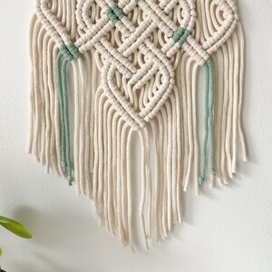May include: A white macrame wall hanging with a diamond pattern and green accents. The hanging has long fringe tassels.