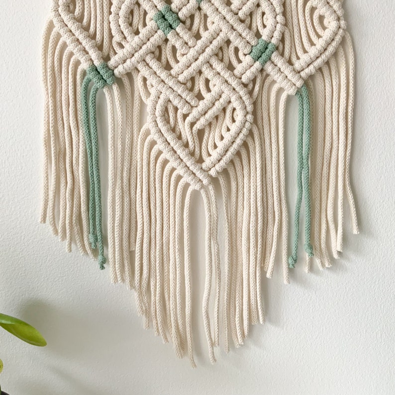 May include: A white macrame wall hanging with a diamond pattern and green accents. The hanging has long fringe tassels.