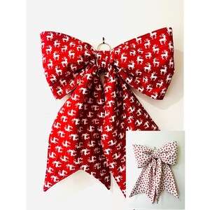 May include: A large fabric bow featuring a vibrant red background with a repeating white pattern of reindeer and thin vertical stripes. The bow includes two long, pointed tails and a central knot with a small metal ring attached for hanging purposes.