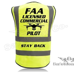 May include: High-visibility yellow safety vest with reflective gray stripes. The vest features black text that reads "FAA LICENSED COMMERCIAL PILOT" with a drone graphic and "STAY BACK". The SafeWear Co. logo is in the bottom right corner.