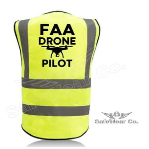 May include: High-visibility yellow safety vest with black text reading "FAA DRONE PILOT" and a drone graphic. The vest has reflective gray stripes and black trim. The SafeWear Co. logo is in the lower right corner.