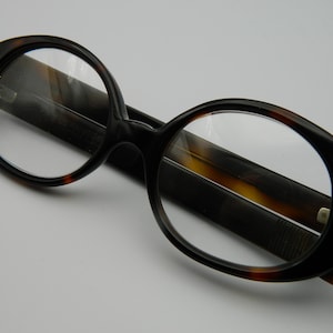 Puede incluir: Gafas de forma ovalada con un patrón marrón oscuro y carey. Las gafas tienen una montura gruesa y lentes transparentes. Las gafas están sobre un estuche marrón oscuro.