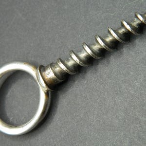 Könnte beinhalten: Ein antiker Metall-Korkenzieher mit einer Wendel und einem kreisförmigen Griff. Der Korkenzieher hat eine silberne Oberfläche mit Gebrauchsspuren, die auf sein Alter hindeuten. Das Design ist einfach und funktional, typisch für Vintage-Barzubehör.