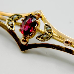 Op de afbeelding: Goudkleurige broche met een ovale, dieprode edelsteen in het midden. De broche heeft delicate blad- en parelaccenten en een gebogen, lineair ontwerp. Het metaal heeft een warme, antieke afwerking.