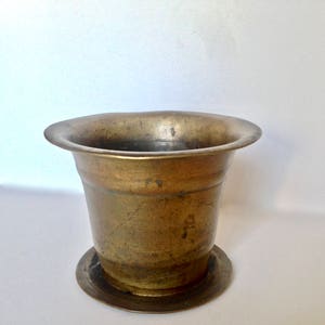 May include: A vintage brass mortar with a wide, flared rim and a circular base. The mortar has a cylindrical body with a slightly tapered shape. The brass has a warm, aged patina, suggesting it is an antique or vintage item. The mortar is likely used for grinding herbs and spices.