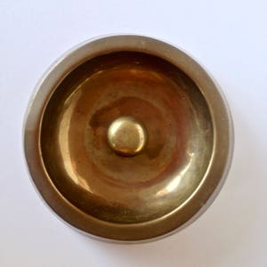 May include: A small, round, brass-colored bowl with a raised center. The bowl has a wide, slightly curved rim and a smooth, polished surface. The interior of the bowl reflects light, creating a warm, golden glow.