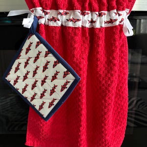 May include: Red kitchen towel with a decorative band featuring red and black fish on a white background. A matching pot holder with the same fish pattern and a navy blue border hangs next to the towel. White ribbons are attached to the towel.