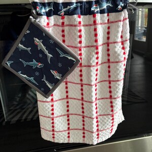 May include: White kitchen towel with red grid pattern and a navy blue shark-themed accent. Includes a matching pot holder with shark print and gray trim. The towel is hanging on a stainless steel oven.