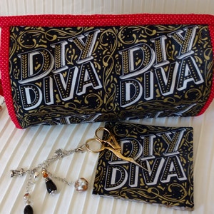 May include: A black and gold fabric makeup bag with a red zipper and white polka dots. The bag has the text "DIY Diva" printed on it. The bag is next to a small black and gold fabric pouch with the same text and a pair of gold scissors.