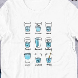 Op de afbeelding: Wit T-shirt met een afbeelding van negen glazen water, elk met een ander persoonlijkheidstype gelabeld, zoals "Optimist", "Pessimist", "Realist", "Fysicus", "Surrealist", "Relativist", "Utopist", "Scepticus" en "Kunstenaar".