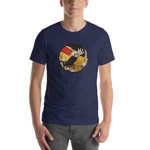 May include: Navy blue t-shirt featuring a circular graphic. The graphic depicts a hand reaching out from a yellow and black background, with a red rectangle and black suit sleeve visible. The shirt is a short-sleeved crew neck.