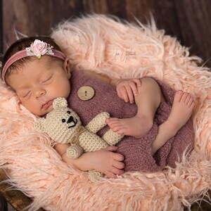 May include: A newborn baby wearing a pink headband with a flower, sleeping peacefully in a wooden bowl lined with fluffy, light pink faux fur. The baby is wrapped in a mauve knit wrap and clutching a small, beige teddy bear.