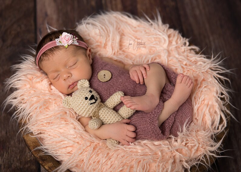 May include: A newborn baby wearing a pink headband with a flower, sleeping peacefully in a wooden bowl lined with fluffy, light pink faux fur. The baby is wrapped in a mauve knit wrap and clutching a small, beige teddy bear.