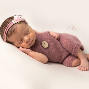 May include: A newborn baby wearing a dusty rose knitted romper with a large wooden button. The baby is also wearing a matching headband with a floral accent. The baby is lying on a cream-colored surface.