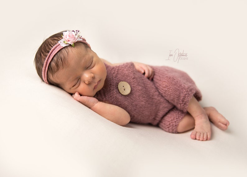 May include: A newborn baby wearing a dusty rose knitted romper with a large wooden button. The baby is also wearing a matching headband with a floral accent. The baby is lying on a cream-colored surface.