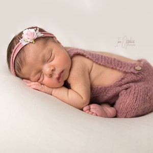 May include: A newborn baby wearing a dusty rose knitted romper with suspenders and a matching headband adorned with a pink flower and white lace. The baby is sleeping peacefully on a cream-colored surface.
