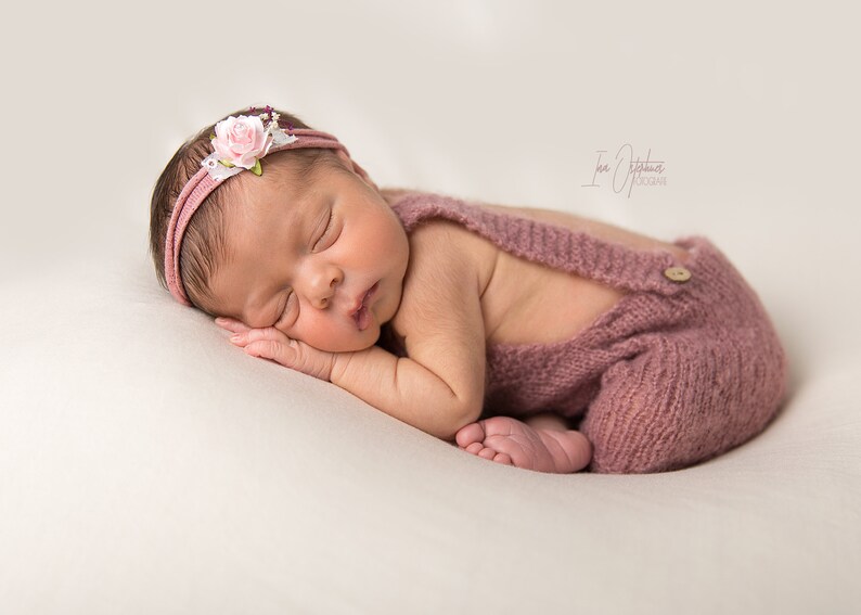 May include: A newborn baby wearing a dusty rose knitted romper with suspenders and a matching headband adorned with a pink flower and white lace. The baby is sleeping peacefully on a cream-colored surface.