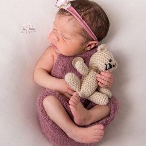 May include: A newborn baby wearing a mauve knitted outfit and a matching headband with a small flower. The baby is holding a small, beige crocheted teddy bear. The baby is lying on a white surface.