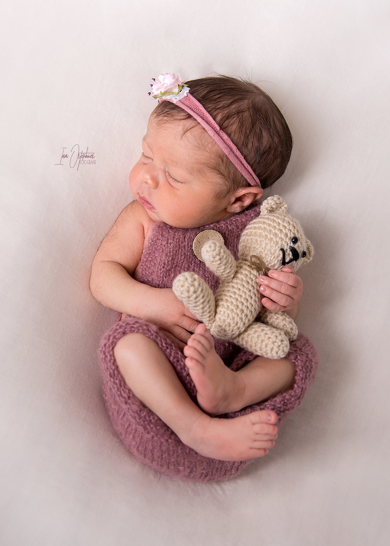 May include: A newborn baby wearing a mauve knitted outfit and a matching headband with a small flower. The baby is holding a small, beige crocheted teddy bear. The baby is lying on a white surface.