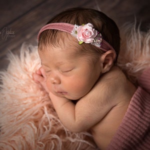 May include: A newborn baby sleeping peacefully, wearing a pink headband with a floral accent. The baby is wrapped in a pink blanket and resting on a fluffy, light pink surface. The background is a wooden surface.