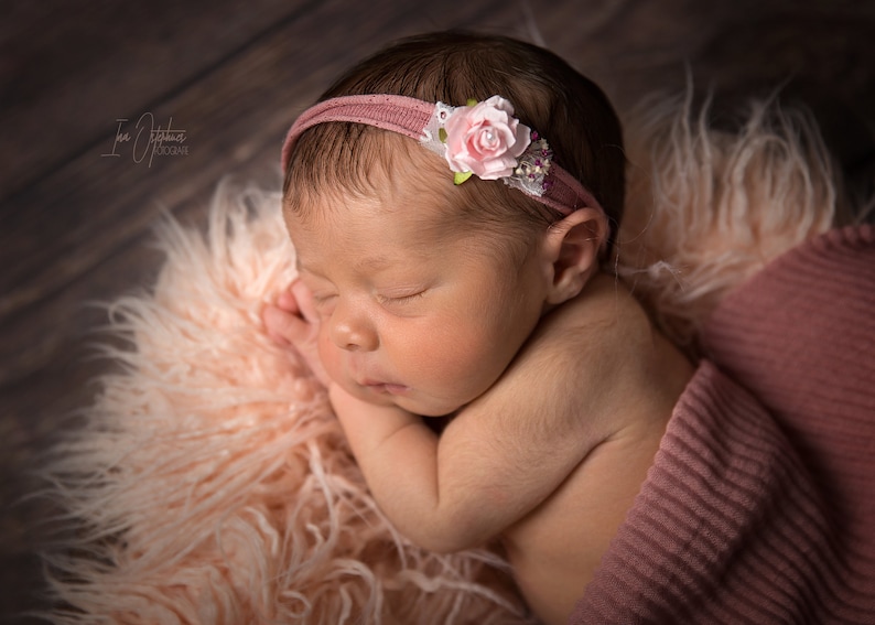 May include: A newborn baby sleeping peacefully, wearing a pink headband with a floral accent. The baby is wrapped in a pink blanket and resting on a fluffy, light pink surface. The background is a wooden surface.