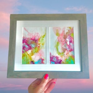 May include: A framed abstract art piece with two panels. The panels feature a swirling, colourful design with shades of pink, green, blue, and purple.