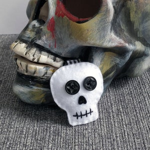 May include: A ceramic skull painted in shades of grey, brown, and yellow with red paint splatters. A small white felt skull with black button eyes and a black stitched mouth is attached to the ceramic skull's jaw.