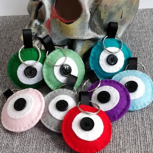 May include: A set of ten felt evil eye keyrings in various colours, including green, teal, purple, pink, grey, and red. Each keyring features a white circle with a black button in the centre, representing the evil eye.