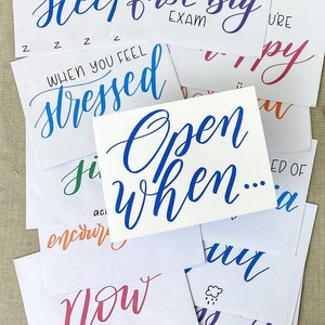 May include: A collection of white cards with handwritten calligraphy in various colors. The cards feature phrases like "Open When...", "When you feel stressed", and "When you take your first big exam". The overall theme is a personalized gift.