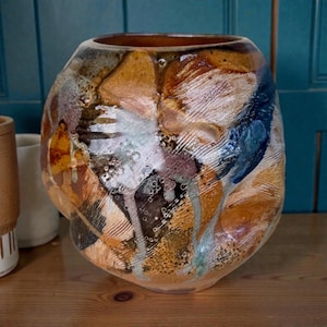 May include: A handcrafted ceramic vase with a unique, organic shape. The vase features a blend of earthy tones, including browns, oranges, and blues, with textured surfaces and dripped glaze effects. The interior is a warm brown.