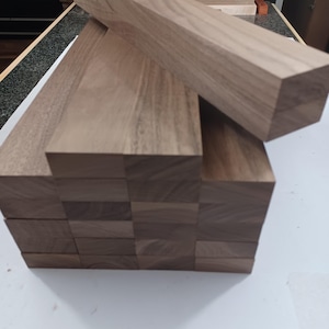 May include: A stack of rectangular brown walnut wood blocks arranged in a grid pattern. The wood features a natural grain texture with varying shades of medium brown. One block is positioned diagonally across the top of the stacked pile.
