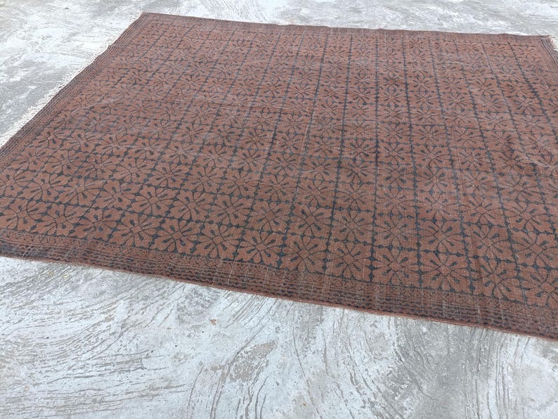 May include: A large, rectangular rug with a repeating floral pattern in dark brown on a lighter brown background. The rug has a border with a similar design. The rug is placed on a concrete surface.